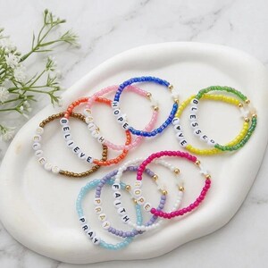 May include: A selection of colourful beaded bracelets, including blue, pink, and green, each with white letter beads spelling out words such as "Believe," "Hope," and "Blessed." The bracelets feature gold-coloured beads and star-shaped accents.