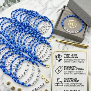 Pode incluir: Uma coleção de pulseiras de contas azuis com letras brancas que formam a palavra PERSIST, contas douradas e pingentes de letras. Inclui uma caixa de presente cinza com logotipo, uma caneta dourada e uma placa com detalhes sobre serviços de marca.