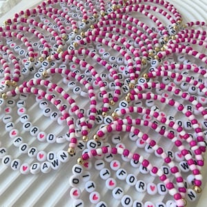 Puede incluir: Una colección de pulseras de cuentas con cuentas circulares blancas con letras negras que forman IT FOR DAWN, intercaladas con cuentas de corazón rosa, cuentas redondas de color rosa vibrante y rosa claro, y pequeñas cuentas doradas.