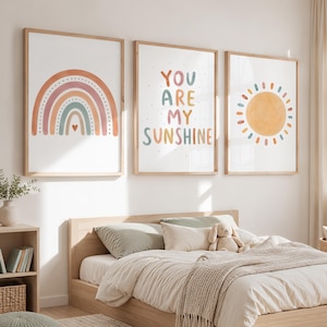 May include: Three framed wall art prints featuring a rainbow, the text YOU ARE MY SUNSHINE, and a sun. The rainbow is composed of muted orange, pink, and blue arches with a small heart. The text is multicoloured, and the sun is yellow.
