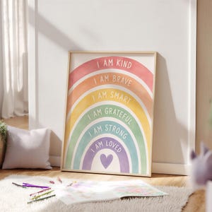 Könnte beinhalten: Ein gerahmter Kunstdruck mit einem Aquarell-Regenbogen in sechs farbigen Streifen. Jeder Streifen enthält weißen Text: I AM KIND, I AM BRAVE, I AM SMART, I AM GRATEFUL, I AM STRONG und I AM LOVED, mit einem kleinen violetten Herz in der Mitte.