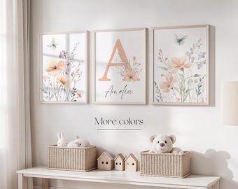 May include: Three framed watercolor prints with a floral theme. The center print features the letter "A" in a peach color with the name "Amelia" written below. The prints are hung on a white wall above a wooden table.