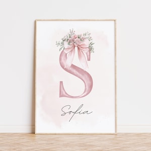May include: A framed watercolor print featuring a large pink letter "S" adorned with a pink bow and floral arrangement. The name "Sofia" is written in cursive below the letter. The background has a soft pink watercolor wash.
