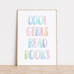 May include: A framed print with the text "COOL GIRLS READ BOOKS" in colorful watercolor lettering. The words are in shades of blue, green, pink, and brown against a white background. The frame is a light wood color.