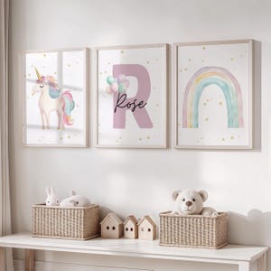 Op de afbeelding: Drie ingelijste kunstdrukken met een eenhoorn, de letter "R" met de naam "Rose" en een regenboog. De eenhoorn heeft een gouden hoorn en een roze en blauwe manen en staart. De letter "R" is roze met ballonnen. De regenboog is in pastelkleuren.