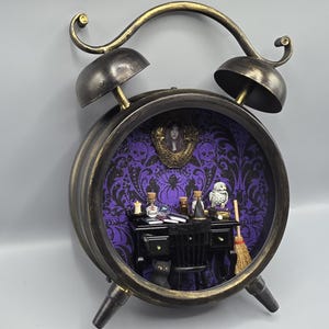 May include: A decorative clock-shaped display case with a black and gold finish. The interior features a purple wallpaper with black skull and spider patterns, a miniature black desk, a chair, a black cat, a broom, and a small white owl.