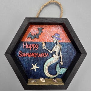 May include: A hexagonal wooden wall hanging with a black frame and jute rope hanger. The background features an orange and blue glitter gradient. It depicts a skeleton mermaid wearing a purple hat, holding a cocktail, with a bat, starfish, and seashells.