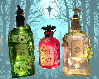May include: Three decorative glass bottles with internal lights. The left bottle is green with a snake label, the center is red with a bat stopper and 'Black Ravens Witch's Drool' label, and the right is clear with a mushroom stopper and 'Nightshade' label.