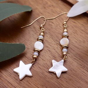 Op de afbeelding: Een paar gouden oorbellen met witte ster- en schelpcharms. De oorbellen hangen aan een houten oppervlak.