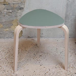 Könnte beinhalten: Ein hellgrüner dreieckiger Hocker mit drei Holzbeinen. Der Hocker ist aus Sperrholz gefertigt und hat eine glatte, lackierte Oberfläche.