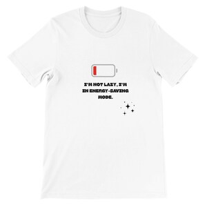 Pode incluir: Camiseta branca com gola redonda e mangas curtas. A camiseta apresenta um gráfico de um ícone de bateria fraca e o texto "I'M NOT LAZY, I'M IN ENERGY-SAVING MODE." com um grupo de estrelas.