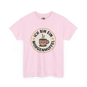 Op de afbeelding: Lichtroze t-shirt met een cirkelvormige afbeelding. De afbeelding bevat de tekst "ICH BIN EIN MORGENMUFFEL" en een cartoon koffiekopje met gesloten ogen. De afbeelding is bruin, beige en wit.