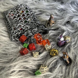 Puede incluir: Una colección de dados poliédricos de varios colores y tamaños, esparcidos sobre una superficie gris y esponjosa. Una bolsa de dados de cota de malla es parcialmente visible, junto con figuras en miniatura. Los dados incluyen dados rojos, naranjas, verdes y blancos.