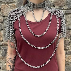 Könnte beinhalten: Ein silbernes Schulterstück aus Kettengeflecht mit passendem Choker und geschichteten Kettenakzenten. Die Metallrüstung wird vor einem bordeauxfarbenen Oberteil und einem Steinhintergrund präsentiert. Das Kettengeflecht besteht aus kleinen, ineinandergreifenden Metallringen.