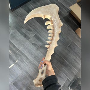 May include: A large, bone-colored prop scythe with a curved blade and a long handle. The handle features a series of tooth-like protrusions along one side. The prop is held in a person's hand.
