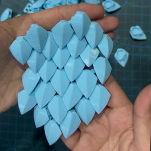 Könnte beinhalten: Eine Ansammlung von hellblauen, 3D-gedruckten Kunststoffschuppen, die in einem flexiblen, ineinandergreifenden Muster angeordnet sind. Jede einzelne Schuppe hat eine spitze, dreieckige Form mit einer texturierten Oberfläche.