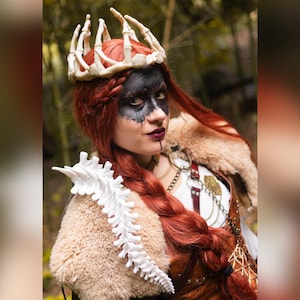May include: A woman with long red hair, wearing a bone crown and a white spine-shaped shoulder piece. She has black face paint and a fur-lined garment. The background is a blurred forest.