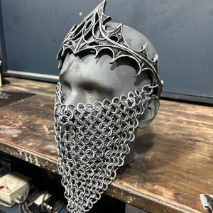 以下が含まれることがあります： 銀色の王冠と鎖かたびらのひげが、灰色のマネキンの頭に展示されています。王冠は装飾的でゴシック様式です。鎖かたびらのひげは、小さな金属リングを組み合わせて作られています。頭は木製の表面にあります。