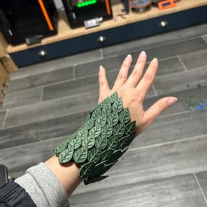 Puede incluir: Un brazalete de color verde oscuro construido a partir de una serie de segmentos superpuestos con forma de hoja. El accesorio presenta una superficie texturizada con venas visibles en cada elemento, creando una apariencia de capas similar a escamas.