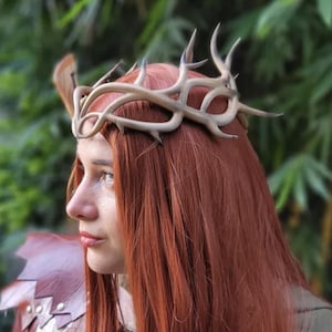 Pode incluir: Uma coroa de chifre castanho-claro com várias pontas. A coroa é usada em cabelo ruivo. O fundo é um verde desfocado. A coroa é um acessório único, perfeito para cosplay ou eventos temáticos.