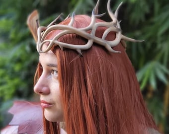 May include: A light brown antler crown with multiple points. The crown is worn on red hair. The background is a blurred green. The crown is a unique accessory, perfect for cosplay or themed events.