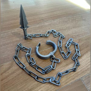 May include: A gray spearhead with a chain and a matching shackle. The spearhead has a pointed tip and serrated edges. The chain is made of interlocking links. The shackle is open and ready to be closed.