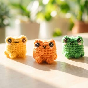 May include: Three small, handmade crochet frogs in yellow, orange, and green. Each frog features large, glossy black safety eyes, a simple stitched mouth, and small blush marks under the eyes. The figures are crafted with a textured yarn stitch.
