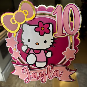Puede incluir: Un adorno para pastel de Hello Kitty rosa y dorado con el número 10. El adorno presenta un diseño de Hello Kitty, estrellas, un lazo y el nombre "Jayla" en cursiva. El diseño está en capas con acentos rosas, dorados y blancos.