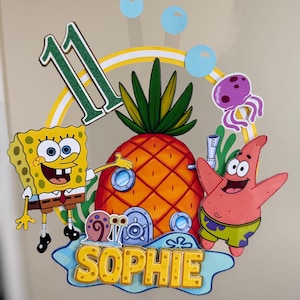 Puede incluir: Un adorno para pastel colorido con Bob Esponja, Patricio Estrella y una casa piña. El número "11" está en verde, y el nombre "Sophie" está en amarillo. Burbujas azules y una medusa morada completan el diseño.