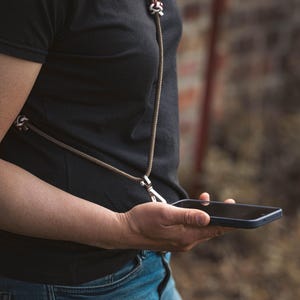 Könnte beinhalten: Ein Handy-Umhängeband aus braunem Seil mit einem silberfarbenen Metall-Drehverschluss und dekorativen Knoten in Weiß und Braun. Die Kordel wird um den Hals getragen und bietet eine sichere Möglichkeit, ein Smartphone zu tragen.