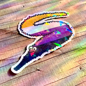 May include: A holographic sticker of a worm with a smiling face. The worm is coloured in a rainbow pattern with yellow, white, purple, and black stripes.
