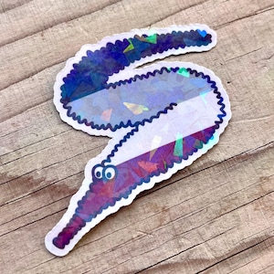 May include: A holographic sticker of a worm with two googly eyes. The worm is a light purple colour with a white stripe down the middle. The sticker is on a wooden surface.