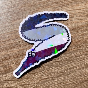 May include: A sticker featuring a whimsical worm design with a holographic finish. The worm is primarily purple, white, and gray, with a black outline and googly eyes. The sticker is on a wooden surface.