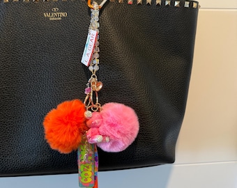Könnte beinhalten: Schwarze Ledertasche mit goldfarbenen Nieten und einem dekorativen Schlüsselanhänger. Der Schlüsselanhänger hat einen orangefarbenen und einen rosa Bommel, einen Anhänger mit dem Text "COZY" und eine Perlenkette mit einem "L.A. GEAR"-Etikett.