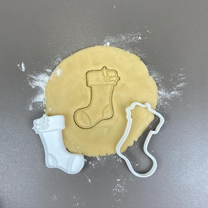 Peut inclure: Un emporte-pièce en plastique blanc en forme de bas de Noël à côté d'une forme de bas découpée dans une pâte à biscuits. Un deuxième emporte-pièce, de type contour, est également présent. De la farine saupoudre la surface grise.