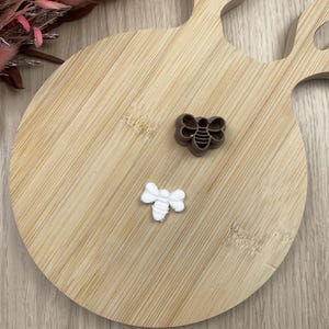May include: A brown bee-shaped cookie cutter and a small white bee-shaped cutout made of clay or dough. The cutter features a detailed outline of a bee with wings and a striped body, while the white cutout shows the final bee shape.