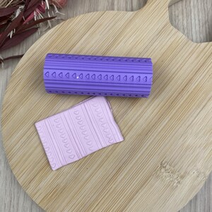 Peut inclure: Un rouleau à texture cylindrique violet orné d'un motif répétitif de petits cœurs et de lignes verticales, accompagné d'un morceau d'argile rectangulaire violet clair présentant le même motif en relief de cœurs et de lignes sur sa surface.