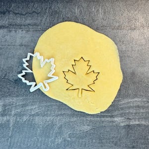 May include: A white plastic maple leaf cookie cutter next to a maple leaf cut-out in a yellow dough. The cookie cutter is on a gray surface. The dough is a light yellow color.