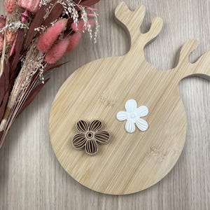 Peut inclure: Une planche à découper en bois clair en forme de tête de renne avec des bois, présentant un emporte-pièce en forme de fleur marron et une pièce découpée en forme de fleur blanche sur sa surface, à côté d'un bouquet de fleurs séchées roses et marron.