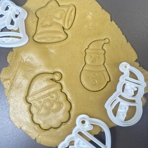 Peut inclure: Gros plan sur de la pâte à biscuits avec des emporte-pièces sur le thème de Noël. La pâte porte les empreintes d'une cloche, d'un bonhomme de neige, du Père Noël et d'un bonhomme de neige. Des emporte-pièces en plastique blanc sont également visibles.