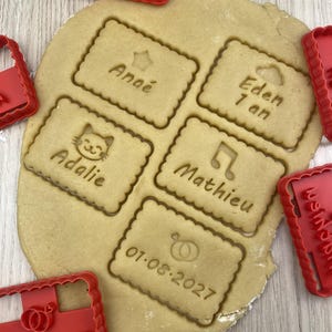 Peut inclure: Cinq empreintes de pâte à biscuits rectangulaires aux bords festonnés, chacune portant un nom ou une date et une petite icône, incluant Anaé, Eden 1 an, Adalie avec un chat, Mathieu avec une note de musique et 01.08.2027 avec des alliances.