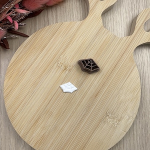 Puede incluir: Un cortador de galletas de plástico marrón con forma de telaraña y una pequeña pieza blanca con forma de telaraña hecha de fondant o arcilla. Los artículos están colocados sobre una tabla de madera clara con forma de cornamenta de ciervo.
