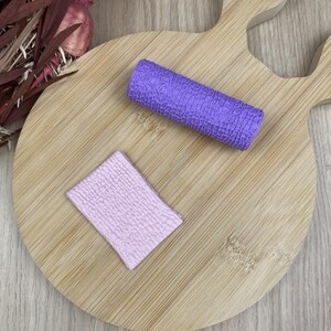 Pode incluir: Um rolo de textura cilíndrico roxo com um padrão de grade detalhado e uma placa de argila retangular roxo-claro com a mesma textura em relevo, ambos concebidos para criar padrões consistentes em materiais de artesanato macios.