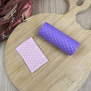 May include: A purple cylindrical roller and a light pink rectangular slab, both featuring a raised, repeating fish scale pattern. The roller and slab are positioned on a light-coloured wooden surface alongside a cluster of dried pink and brown foliage.