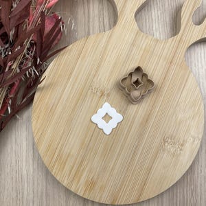 Peut inclure: Un emporte-pièce en plastique beige en forme d'étoile à quatre branches avec un bord festonné, accompagné d'une découpe en argile blanche de la même forme, présentant un vide central en forme de losange dans ce design floral symétrique.