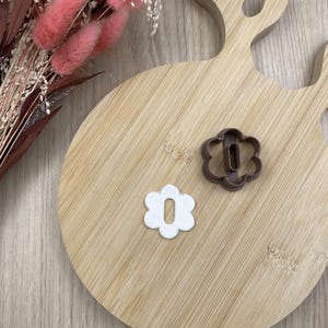 以下が含まれることがあります： 中央に楕円形の切り抜きがある茶色のプラスチック製花型クッキーカッターと、中央に楕円形の穴が開いた白い粘土製の花型が並んでおり、どちらも6枚の丸みを帯びた花びらを持ち、明るい木製の表面に置かれています。