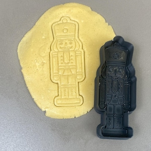 May include: A dark grey plastic cookie cutter shaped like a nutcracker soldier is next to a piece of dough with the nutcracker shape pressed into it. The dough is light yellow.