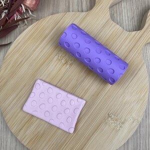 Peut inclure: Un rouleau à pâtisserie cylindrique violet avec un motif de cercles en relief répété et une plaque rectangulaire en matériau violet clair présentant le même motif de cercles en relief, avec une texture uniforme sur les surfaces.