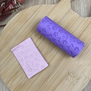 Peut inclure: Un rouleau à pâtisserie cylindrique violet avec un motif en relief répété d'empreintes de pattes de chien et d'os, accompagné d'un morceau rectangulaire d'argile rose clair présentant le même motif imprimé de pattes et d'os.