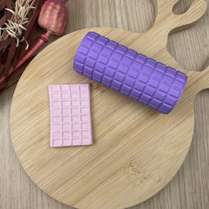 May include: A lilac foam roller and a pale pink foam block with a grid pattern are displayed on a wooden cutting board. The cutting board has a deer head shape. Dried flowers and a seed pod are in the background.